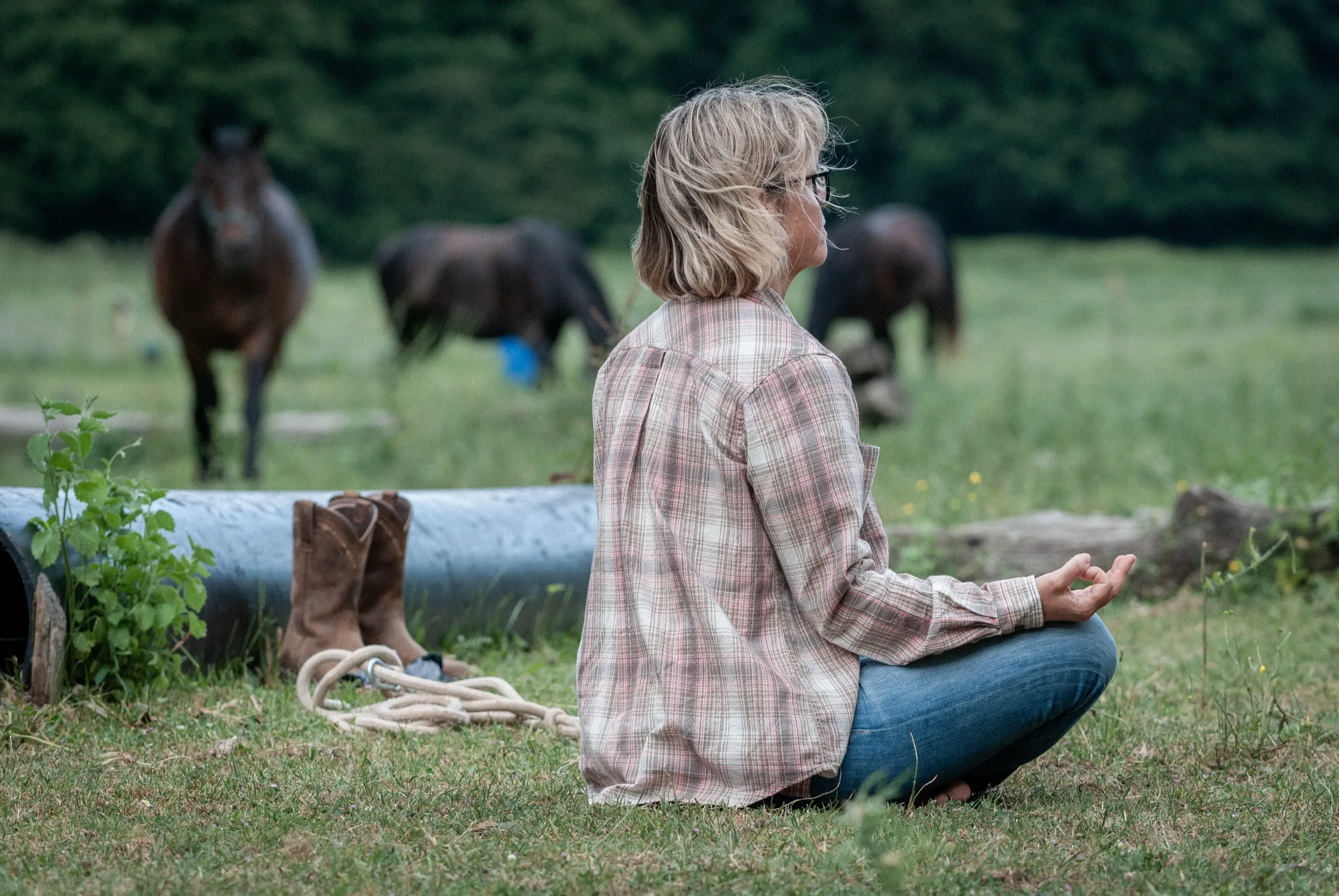 A woman sitting in quiet meditation as horses graze behind her