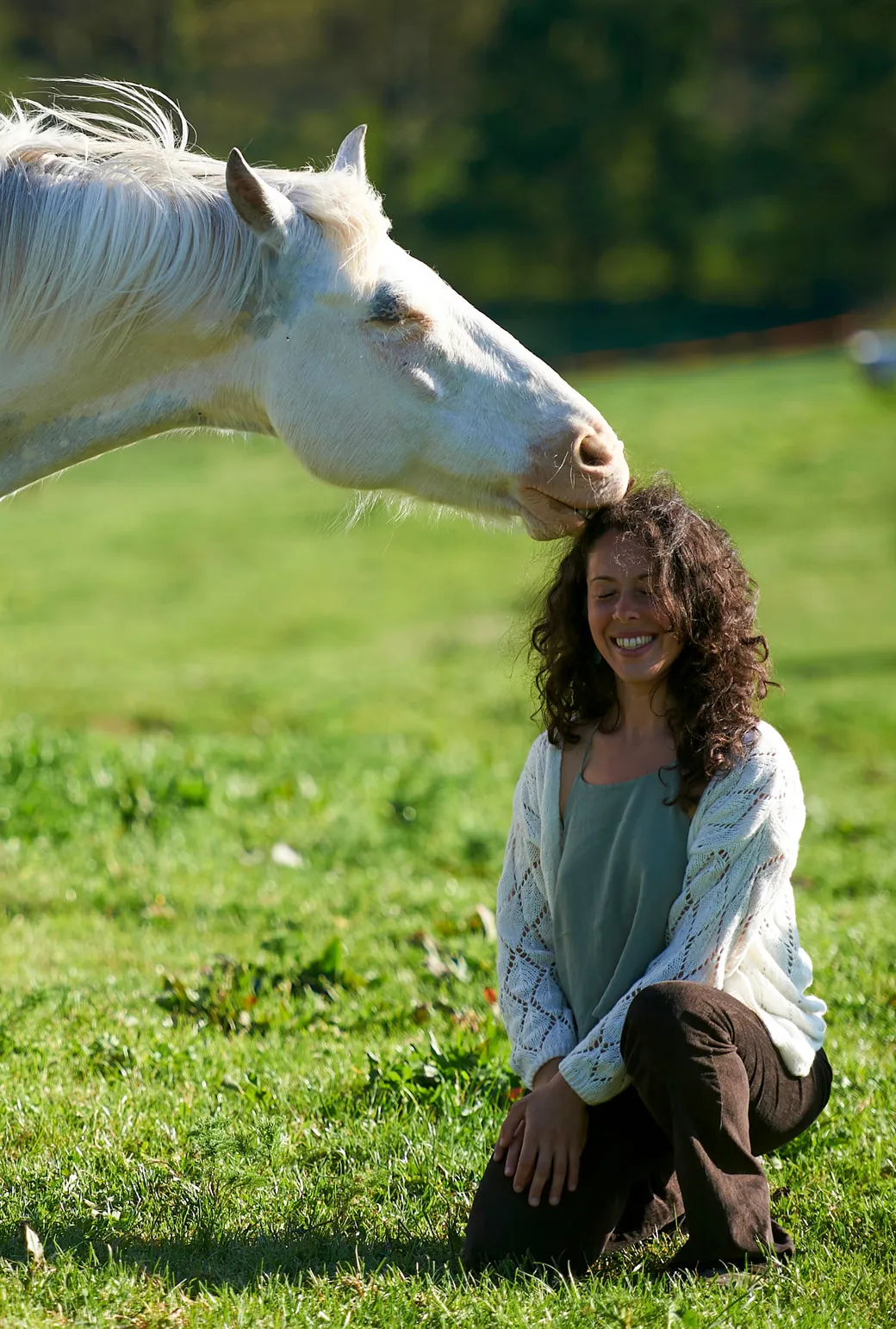 A white horse lowering its muzzle to the head of a kneeling, smiling woman in a sunlit meadow