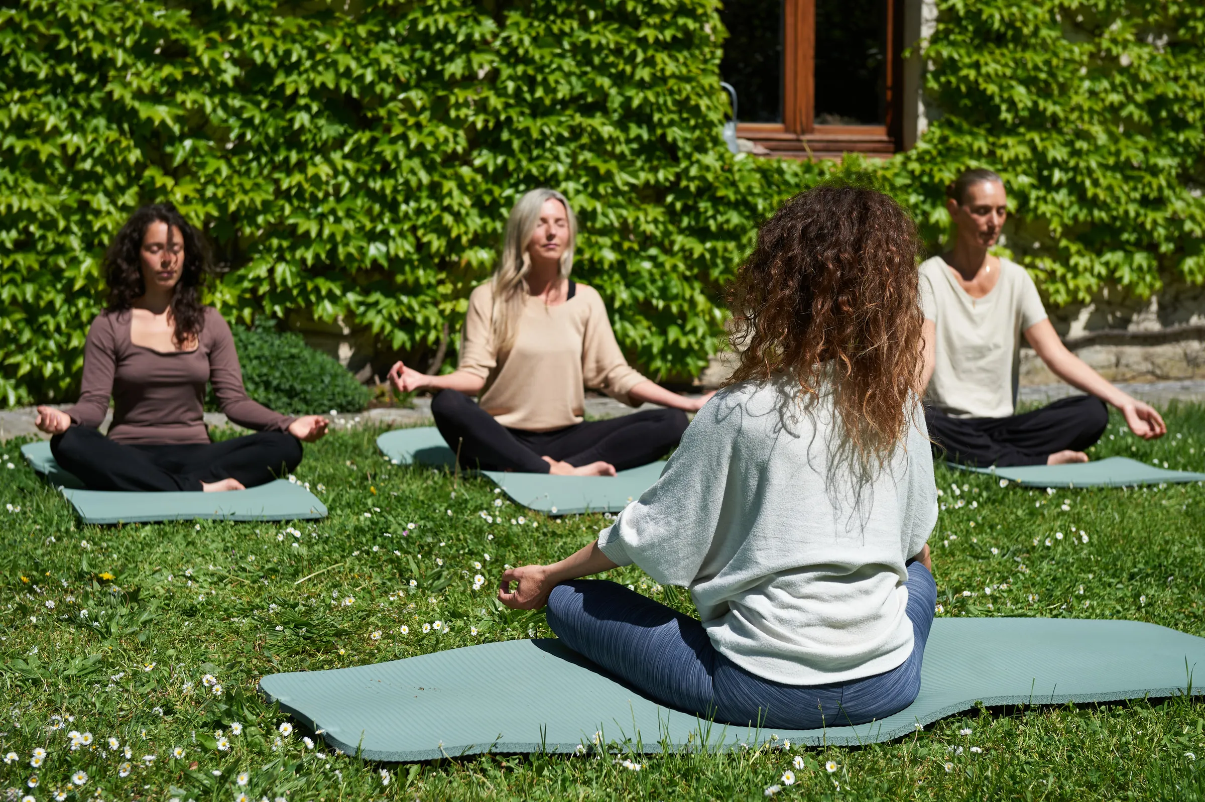 Four women sitting cross-legged in meditation on yoga mats on a lawn beside an ivy-covered wall