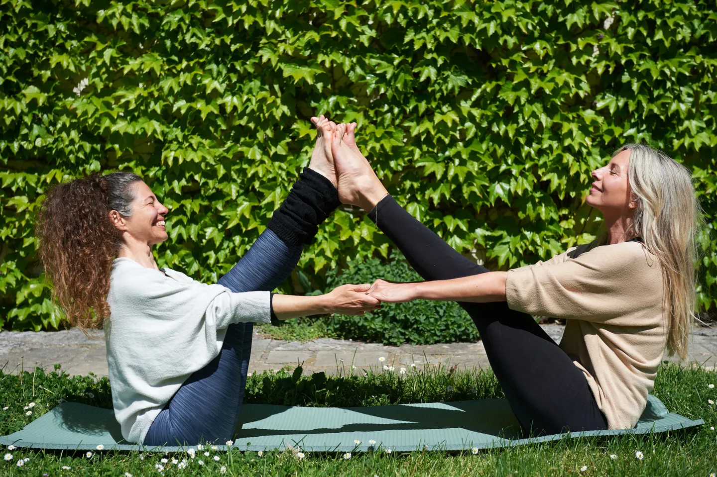 Two women practising a partner yoga pose together on mats on the lawn, hands and feet joined